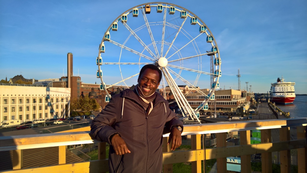 Kuvassa on Olumide Aluko. Kuva on otettu ulkona ja taustalla näkyy maailmanpyörä. Alumidella on tummat vaatteet ja hän nojaa kaiteeseen. Hän hymyilee kuvassa.