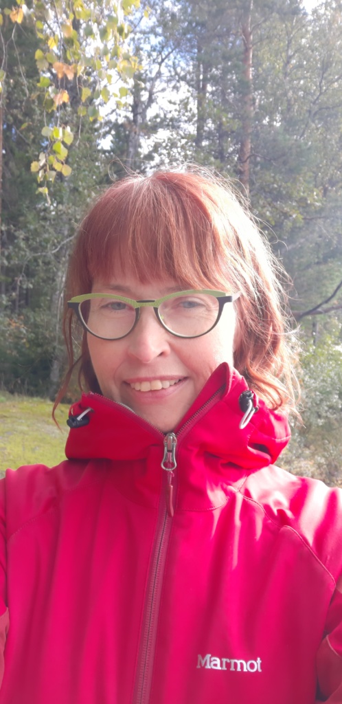 Kuvassa Sari Ruottinen. Sarilla on kuvassa hiukset auki. Sarilla on silmälasit ja pinkki takki. Kuva on otettu ulkona. Sari hymyilee kuvassa.